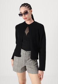 Μαύρο πλεκτό ζακέτα με στρογγυλή λαιμόκοψη, που διαθέτει κούμπωμα με ένα κουμπί. Συνδυάζεται με γκρι καρό σορτς και μαύρο τοπ.