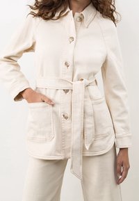 Beige canvasjacka med knapprad, bältad midja och stora påsydda fickor. Har en krage och synliga sömnadsdetaljer.