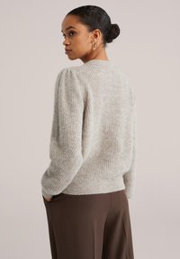 Světlé šedý pletený svetr s texturovaným povrchem, kulatým výstřihem a mírně nafouklými rukávy. Nošený s hnědými kalhotami.