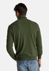 Jersey de cuello alto de punto en verde oscuro, con puños y dobladillo acanalados. Presenta una textura suave y un diseño ajustado, lucido sobre unos jeans de color azul claro.