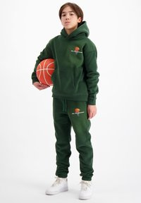 Donkergroene hoodie met kangoeroezak en geborduurd logo, gecombineerd met bijpassende joggers. Witte basketbal tegen het lichaam gehouden; witte sneakers.