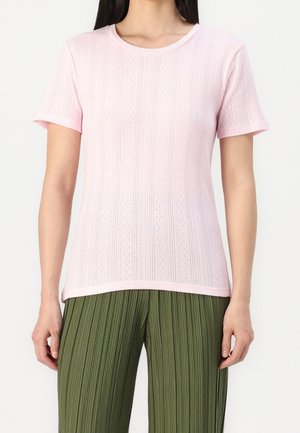 Nainen, joka käyttää vaaleanpunaista lyhythihaista neulepuseroa, jossa on hienovaraisia pystysuoria kuvioita, ja oliivinvihreitä laskostettuja housuja. Hän seisoo valkoista taustaa vasten.