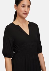 Schwarzes Kleid aus weichem Stoff, mit V-Ausschnitt, Puffärmeln und gerafftem Taillenbereich für zusätzlichen Schwung und Textur.