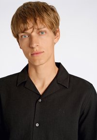 Chemise noire à manches longues avec col court, tissu texturé, broderie de logo discrète sur la poitrine et fermeture à boutons classique.
