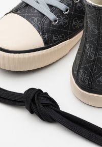 Zapatillas negras con un patrón, con una puntera de goma beige y una suela texturizada. Cuerdas grises y un cordón negro anudado están cerca.