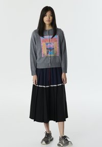 Cardigan gris avec un design imprimé coloré associé à une jupe plissée bleu marine ornée de dentelle et des baskets noires.