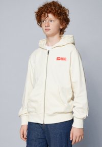 Felpa crema con zip, tasca a marsupio, polsini elasticizzati e logo rosso Diesel sul petto. Tessuto morbido con una vestibilità ampia.