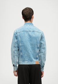 Άνδρας στραμμένος μακριά φορώντας μια ανοιχτόχρωμη μπλε τζιν ζακέτα και μαύρο παντελόνι σε ένα απλό λευκό φόντο.