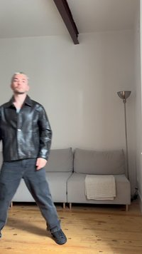 Junger Mann mit blondem Haar trägt eine schwarze Lederjacke und dunkle Hose und steht in einem spärlich eingerichteten Zimmer mit einem grauen Sofa und einer Stehlampe.