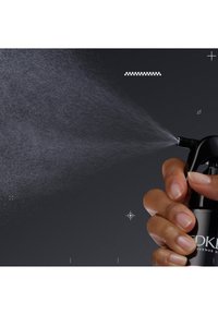 Svart sprayflaska som hålls i en hand, med en fin dimma som sprutas ut. Munstycket är vinklat, och sprayen producerar en delikat, jämn dimma av vätska.