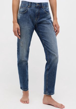 Vrouw draagt mid-rise slim-fit blauwe spijkerbroek met lichte slijtage op dijen en kuiten, steekt haar voeten bloot en staat tegen een effen achtergrond.