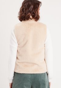 Gilet en polaire avec un dos beige et des manches blanches, présentant une texture douce. Le bas est arrondi et le tissu semble duveteux.