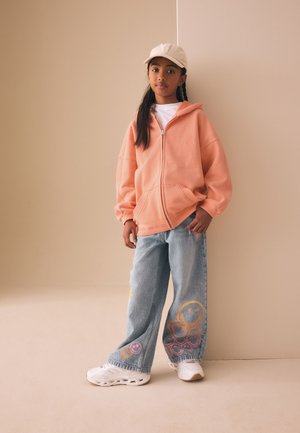 Ung pige står mod en beige væg iført en ferskenfarvet hættetrøje, hvid kasket, vide jeans med farverige smiley-ansigter og hvide sneakers.