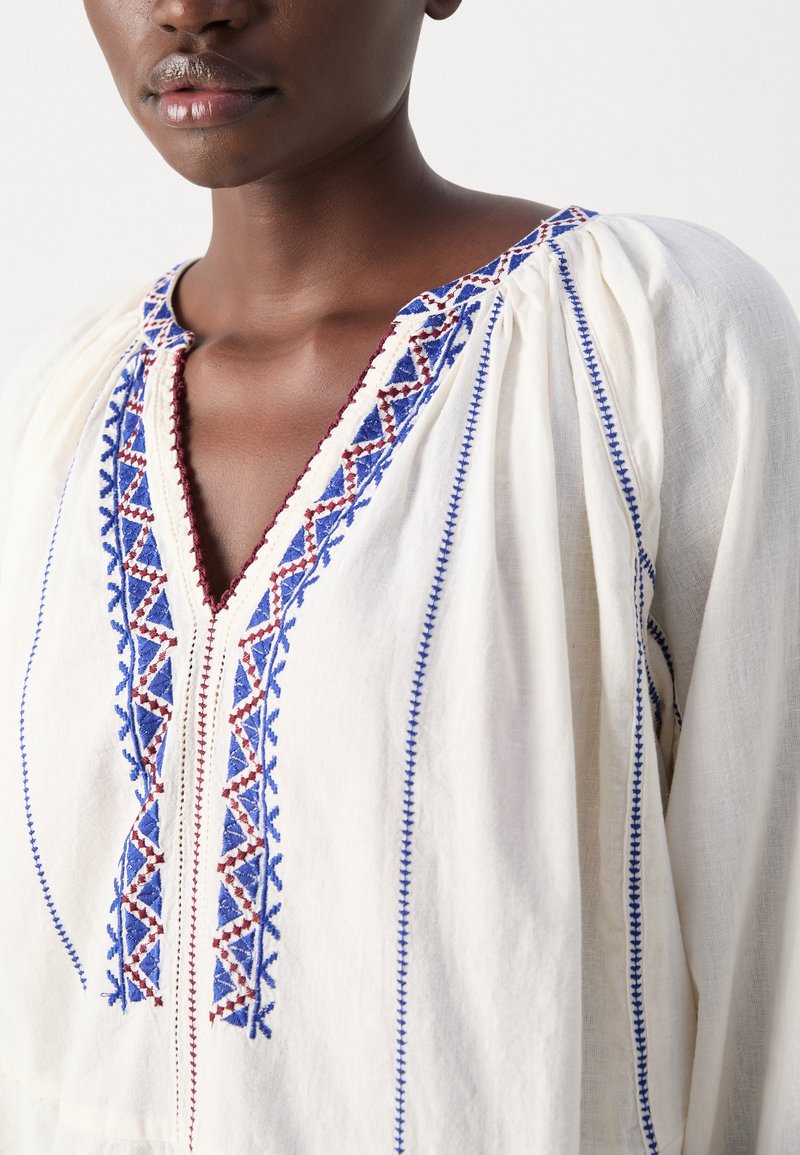 Blouse blanche ornée de motifs brodés bleu et bourgogne le long de l'encolure et de la patte de boutonnage, avec un tissu texturé et bouffant.