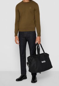 Pull tricot vert olive avec poignets côtelés, jean foncé, et sac en nylon noir avec le texte blanc "BOSS", porté par des anses noires.