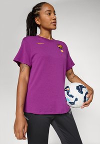 Ljubičasta majica kratkih rukava s Nike logom i grbom Barcelone, u paru s crnim hlačama. Osoba drži loptu za nogomet s uzorkom.