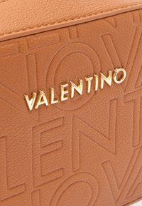Nærbilde av lysebrunt teksturert skinn med preget Valentino-logo og gullfarget metallisk Valentino-bokstaver på et luksuriøst tilbehør.