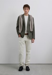 Conjunto masculino que incluye un blazer de espiga gris, un suéter crema, pantalones blancos y zapatos negros, con una bufanda de punto gris caída sobre los hombros.