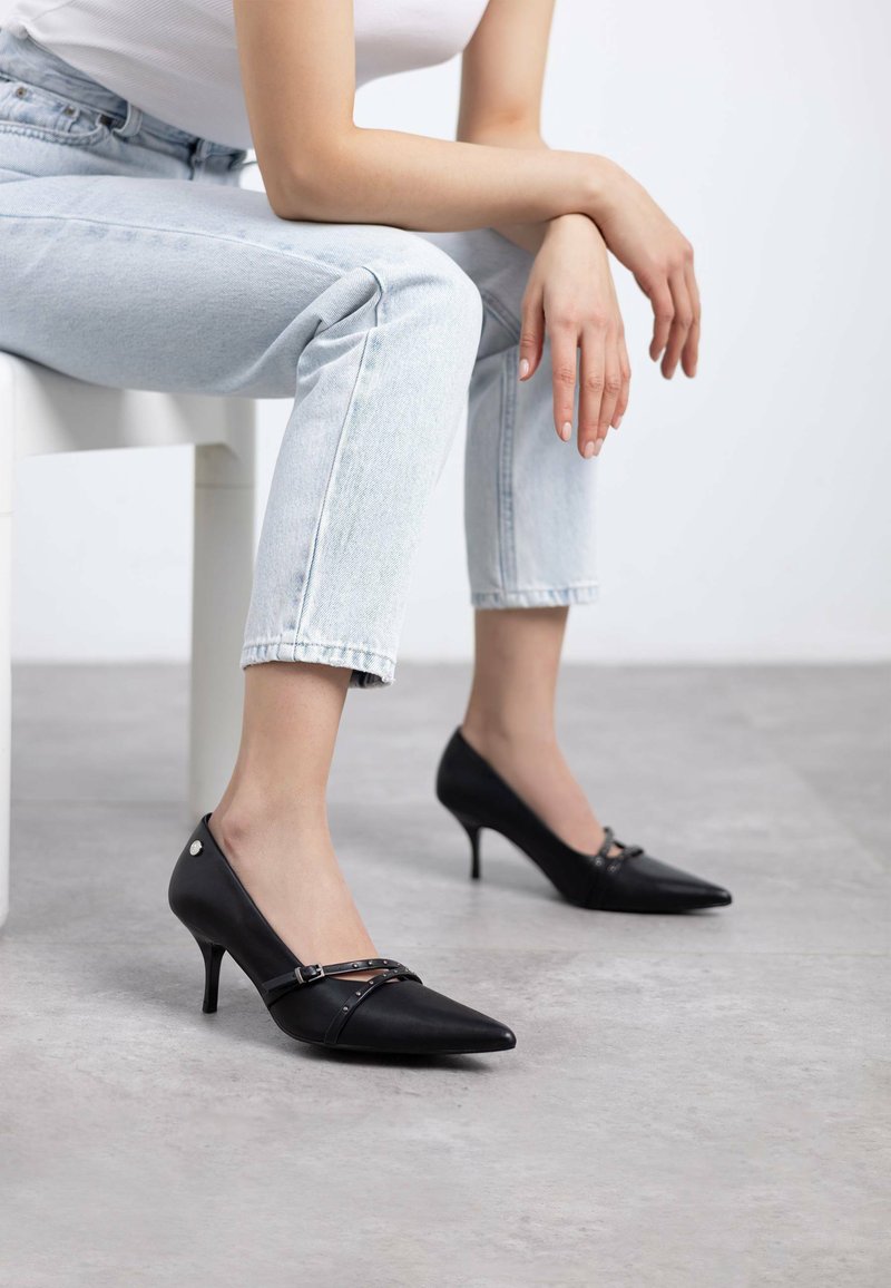 Tacchi alti in pelle nera con punte affusolate, sottili tacchi a stiletto e cinturino decorativo con borchie. Indossati con denim blu chiaro.