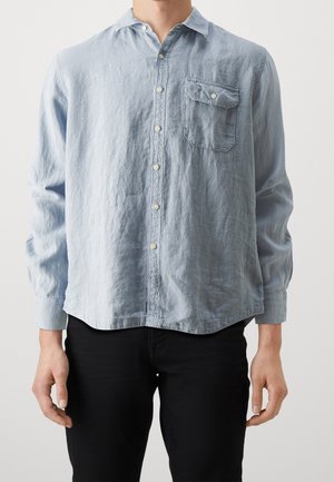 Hombre vistiendo una camiseta de botones de manga larga azul claro con un bolsillo en el pecho abotonado y pantalones negros, contra un fondo blanco liso.