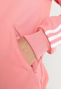 Rosa zip-up sweatshirt med sidoficka och ribbade ärmslut, prydd med tre vita ränder på ärmarna. Mjuk och slät tygkänsla.