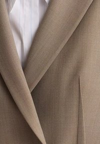 Gros plan sur le revers d'un blazer beige porté sur une chemise blanche à fines rayures bleues, montrant la texture du tissu et le détail de la poche cintrée.