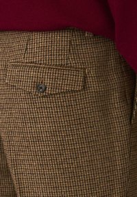 Brown check-patterned trousers with a flat back pocket and a black button closure, made from textured fabric.