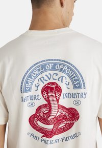 Camiseta de algodón color crema con un gran gráfico de una serpiente roja y texto azul que dice "Equilibrio de Opuestos", "Industria Natural" y "Pasado-Presente-Futuro".