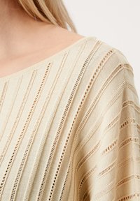 Beige pletený top s vertikálními pruhy, otevřenými pletenými sekcemi a širokým výstřihem, vyrobený z jemné, lehké látky.