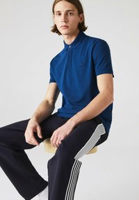 Marineblå pikétröja med korta ärmar och krage, med en diskret logotyp. Matchad med mörka byxor som har vita sidostråk.