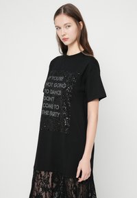 Juoda trumpomis rankovėmis marškinėliai su stačiakampiu blizgučių piešiniu, kuriame pateiktas tekstas. Marškinėliai yra laisvo kirpimo ir turi lygią tekstūrą.