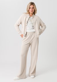 Beige ritsjack gemaakt van zachte stof, gecombineerd met een wit shirt met gouden lijntekeningen, en bijpassende wijde broek in een lichte tint.