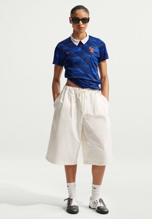 Femeie purtând tricou polo albastru cu model și guler alb, pantaloni scurți albi supradimensionați, ochelari de soare, șosete albe și pantofi negri cu alb, stând cu mâinile în buzunare.