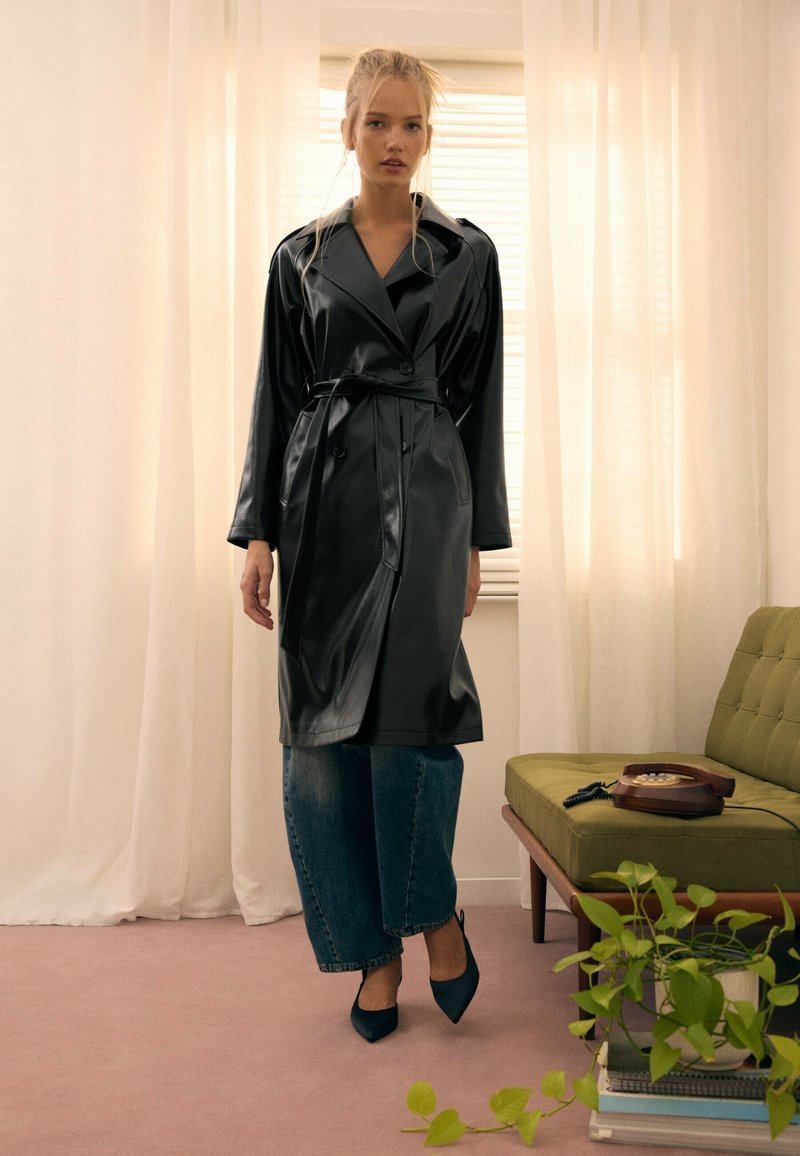 Jeune femme portant un trench-coat en cuir noir, un jean large bleu et des talons noirs, debout dans une pièce doucement éclairée avec un canapé vert et une plante en pot.