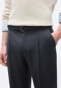 Άνδρας που φορά μαύρο παντελόνι ριγέ, μαύρη ζώνη, κρεμ πλεκτό πουλόβερ και μπλε ριγέ πουκάμισο με τα μανίκια ανασηκωμένα.