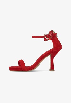 Czerwona zamszowa sandałka na wysokim obcasie typu słupek, z paskiem na przodzie i paskiem wokół kostki ozdobionym dekoracyjną klamrą z biżuterią.