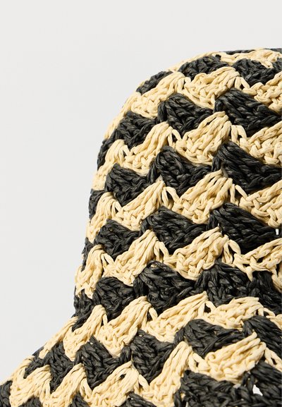 Πλεκτό καπέλο από άχυρο παρουσιάζει μοτίβο ζιγκ-ζαγκ σε μαύρο και μπεζ, με υφασμένη υφή και καμπυλωτό γείσο. Ανθεκτικό υλικό με λεπτομερείς ραφές.