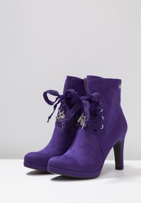 Un par de botines de tacón alto de ante morado con lazos de encaje decorativos y encantos plateados en los cordones, exhibidos sobre una superficie blanca.