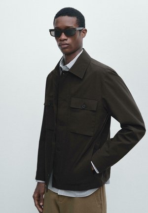 Mand iført mørkebrun jakke med frontlommer med klap, stribet skjorte, kaki bukser og sorte rektangulære solbriller foran en ensfarvet baggrund.