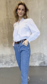 Hellblau-weiß gestreiftes Hemd mit Rüschenkragen, kombiniert mit blauen Jeans und einem braunen Gürtel. Das Model posiert mit den Händen in der Hüfte.