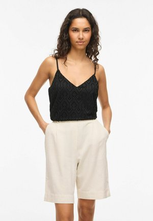 Ung kvinne med krøllete hår iført en svart blondetopp og beige shorts med høy midje, hender i lommer, nøytral bakgrunn.