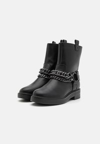 Černé kotníkové boty mají elegantní design se zdobným stříbrným řetízkem a sponou kolem kotníku, umístěné na jednoduchém bílém pozadí.