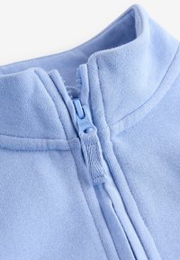 Gros plan du col d'une veste polaire bleu clair avec une fermeture éclair en plastique partiellement fermée et une languette en tissu.