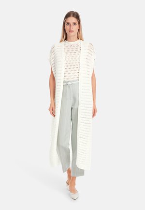 Weißer, offener Strick-Langcardigan über einem weißen, gemusterten Oberteil, kombiniert mit hellgrauen, gestreiften Hosen und spitzen Absatzschuhen. Glatte Textur.