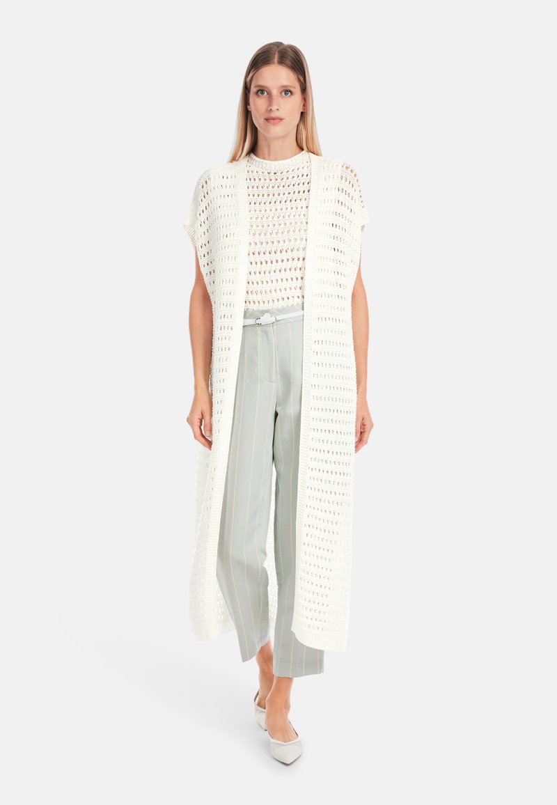 Weißer, offener Strick-Langcardigan über einem weißen, gemusterten Oberteil, kombiniert mit hellgrauen, gestreiften Hosen und spitzen Absatzschuhen. Glatte Textur.