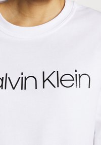 Nära bild av en person som bär en vit t-shirt med rund hals och "Calvin Klein" tryckt i svart över bröstet.