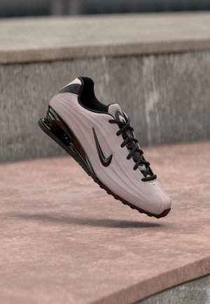 Világos rózsaszín és fekete Nike sportcipő fényes fekete talppal, fekete fűzőkkel és logóval, amely egy betonfelület és fal háttere fölött lebeg.