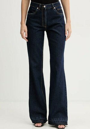 Jeans blu scuro a vita alta con gamba larga, tasche frontali e cuciture visibili, indossati con tacchi neri aperti in punta.