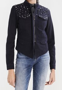 Marinblå jacka med nitar på axlarna, framzip och två bröstfickor, med en strukturerad design och texturerad tyg.