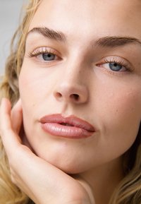 Primo piano di un volto con pelle chiara, occhi azzurri chiari e trucco naturale delicato; presenta sopracciglia ben definite e labbra lisce e lucide.